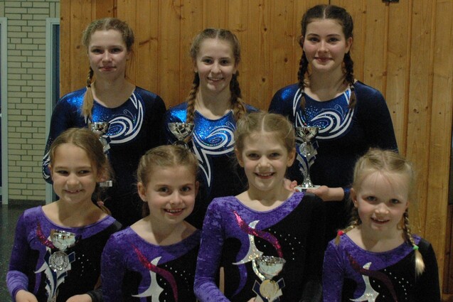 Gruppe von sieben Turnerinnen in Bodys, l&auml;chelnd mit Pokalen, vor einer Holzwand in einer Sporthalle.