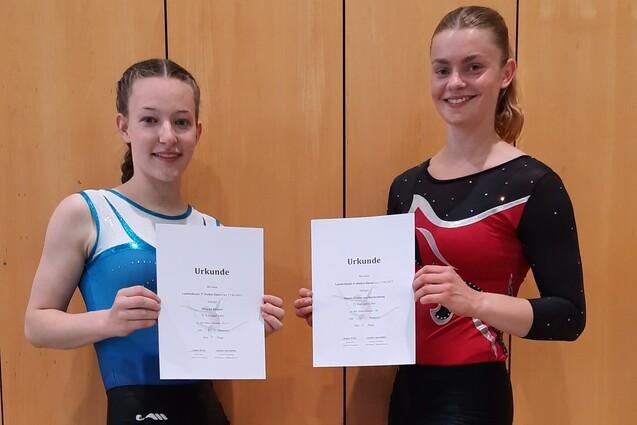 Zwei Turnerinnen in Wettkampfanz&uuml;gen halten stolz ihre Urkunden vor einer Holzpaneelwand.