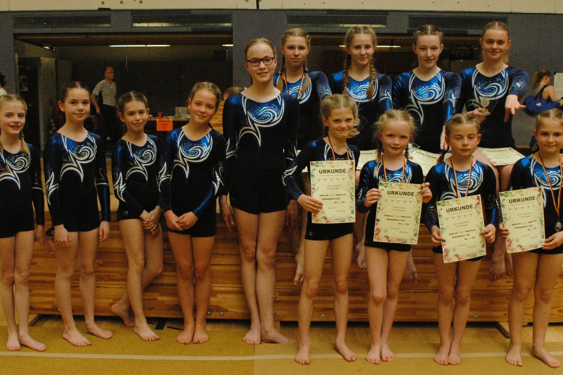 Gruppe von elf jungen Turnerinnen in Wettkampfanz&uuml;gen mit Urkunden auf einem Turnboden in einer Sporthalle.