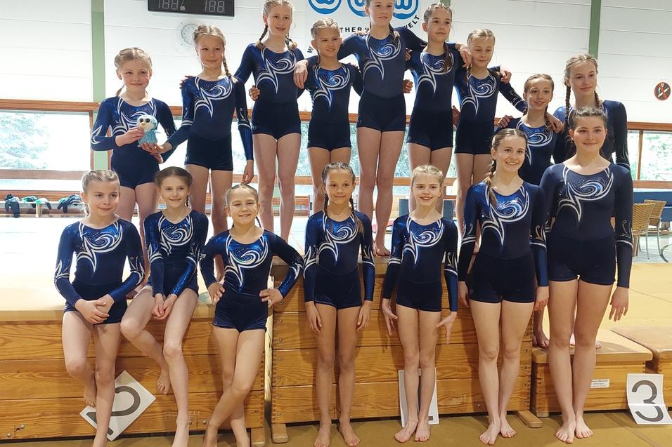 Gruppe von Turnerinnen in blauen Einteileranz&uuml;gen, l&auml;chelnd auf einem Podest im Gymnastikraum.