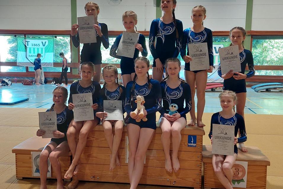 Gruppe junger Turnerinnen in blauen Trikots posiert mit Urkunden auf einem Sprungkasten in einer Turnhalle.