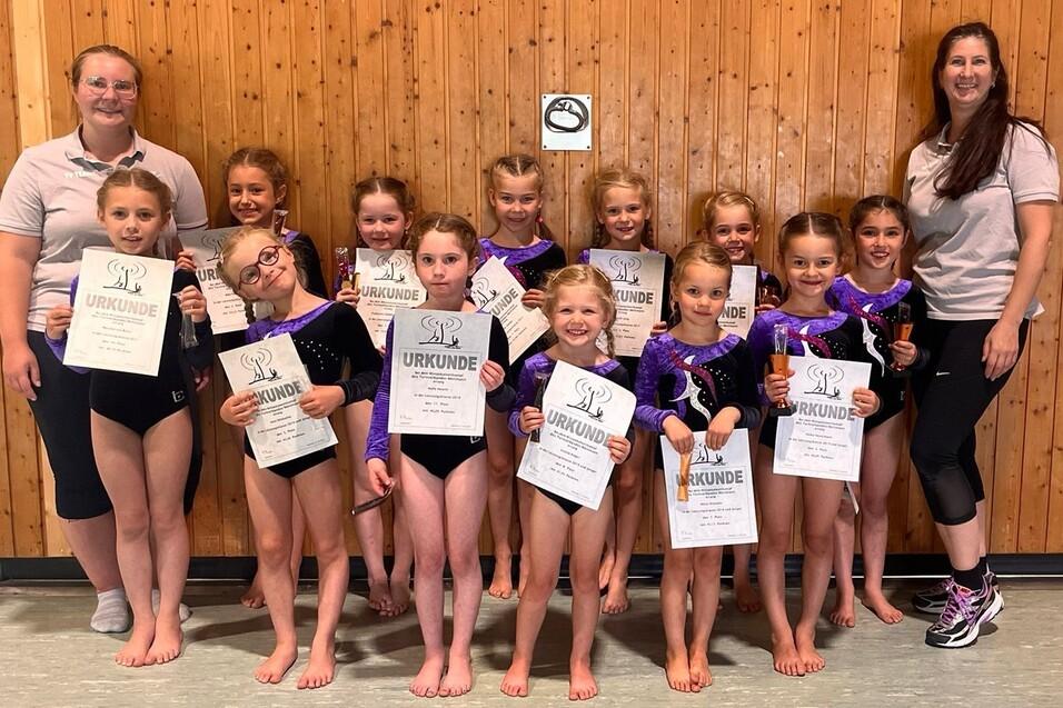 Gruppe von Kindern in Gymnastikanz&uuml;gen h&auml;lt Urkunden, l&auml;chelt vor einer Holzwand mit zwei Trainerinnen.