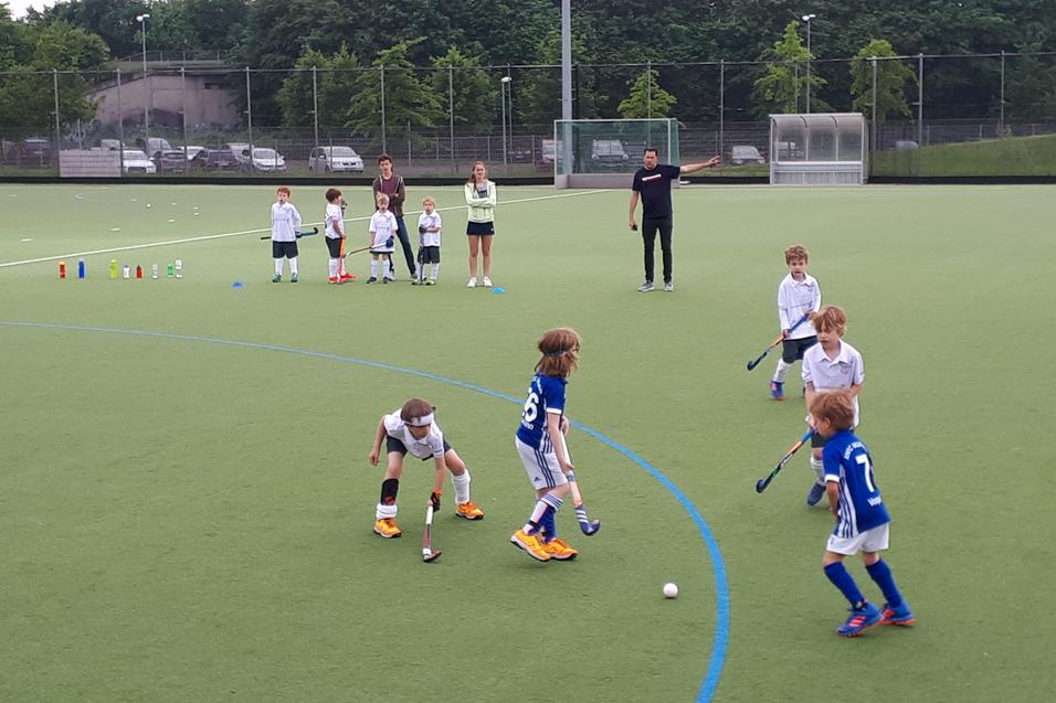 Kinder spielen Hockey auf einem Kunstrasenplatz, w&auml;hrend Trainer sie anleiten. Wasserflaschen stehen bereit.