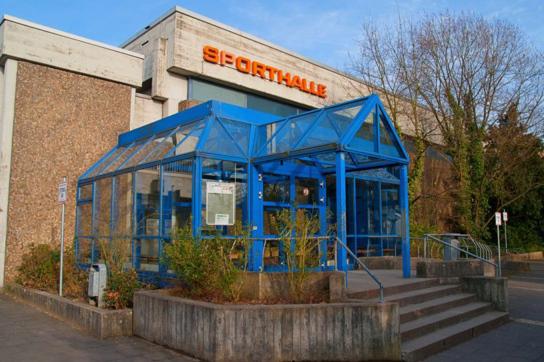 Eingang der Sporthalle mit blauer Glasarchitektur und Pflanzen, beschriftet mit dem Wort &bdquo;SPORTHALLE&ldquo;.