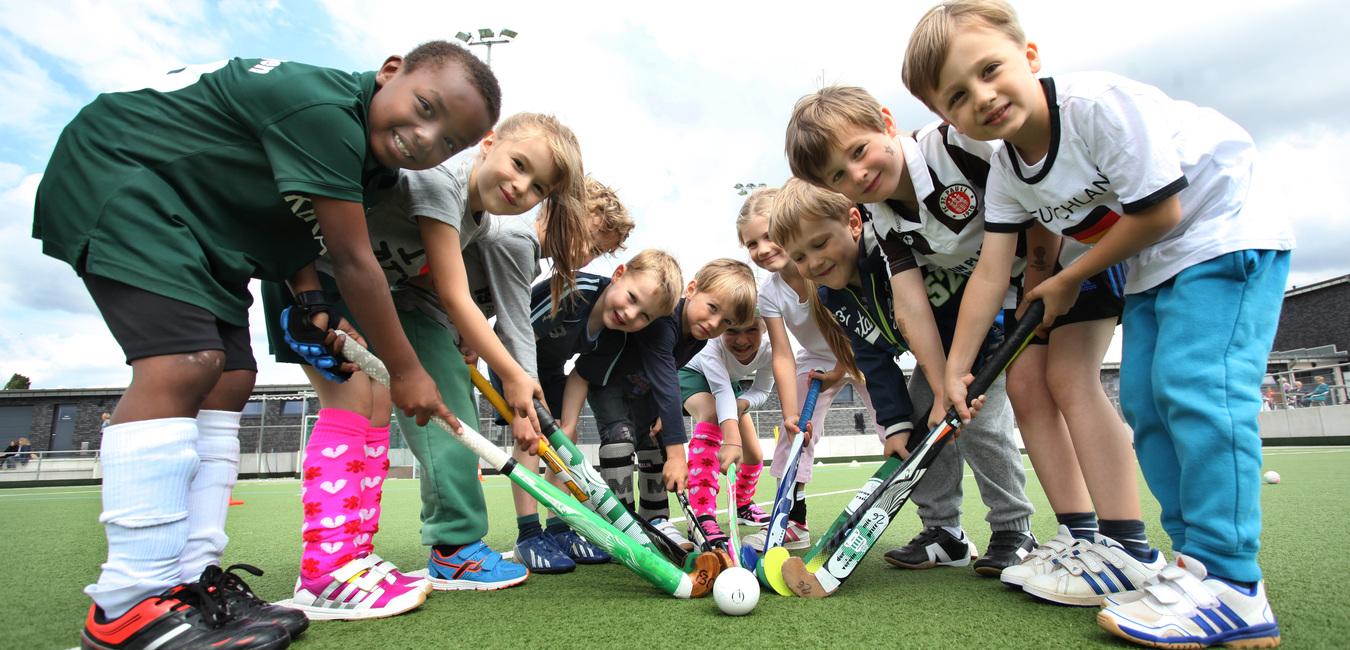 Gruppierung von Kindern in Sportkleidung, die gemeinsam Schl&auml;ger um einen Hockeyball auf einem Kunstrasenplatz halten.