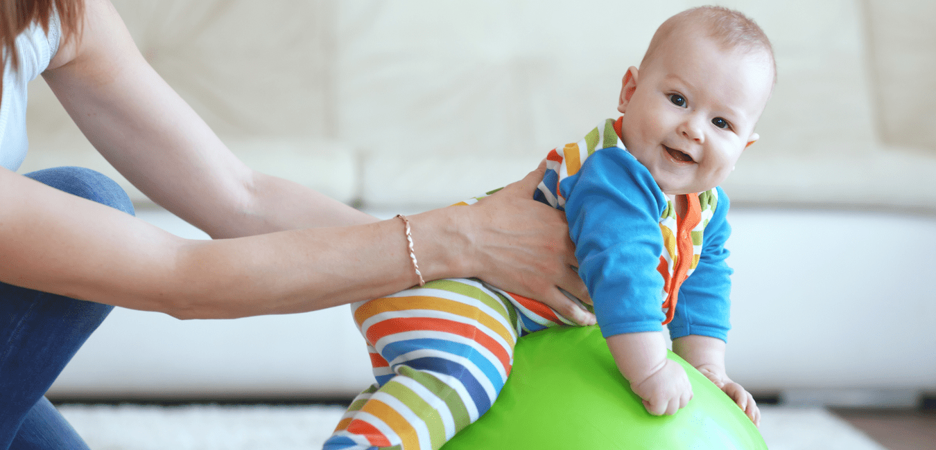 Baby im bunten Strampler sitzt auf einem gr&uuml;nen Gymnastikball, w&auml;hrend eine Hand es sanft st&uuml;tzt.