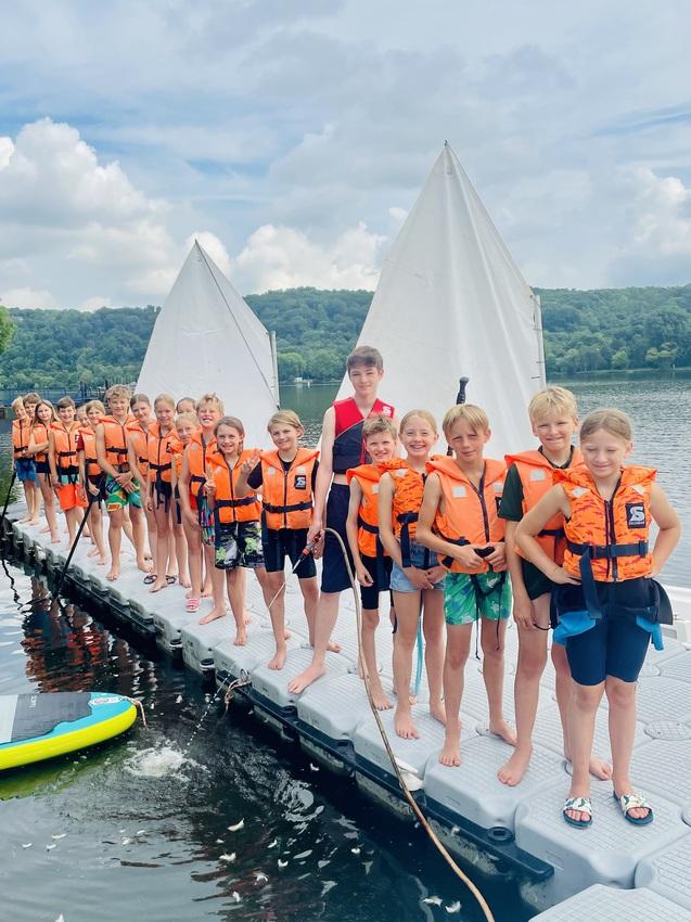 Gruppe von Kindern in Schwimmwesten steht auf einem Steg vor einem See, mit Segelbooten im Hintergrund.