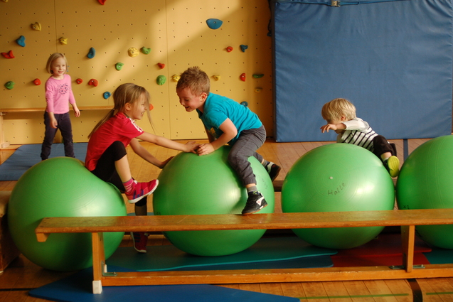Vier Kinder spielen in einer Turnhalle auf gro&szlig;en gr&uuml;nen Gymnastikb&auml;llen nahe einer Kletterwand.