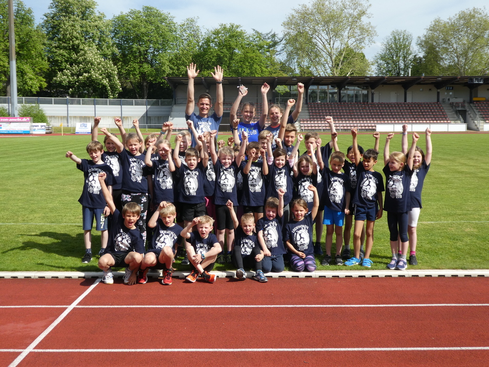 Gruppe von Kindern in T-Shirts mit Aufdruck stehen auf einem Sportplatz, heben jubelnd die Arme in die Luft.