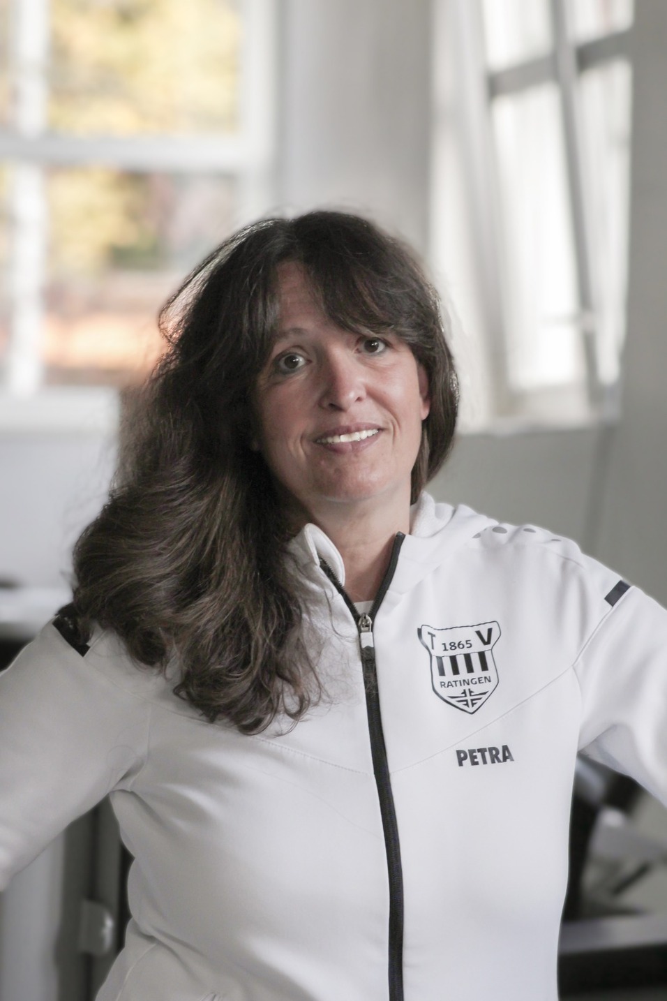 Frau mit langen, lockigen Haaren tr&auml;gt eine wei&szlig;e Jacke mit Vereinslogo und blickt l&auml;chelnd in die Kamera.