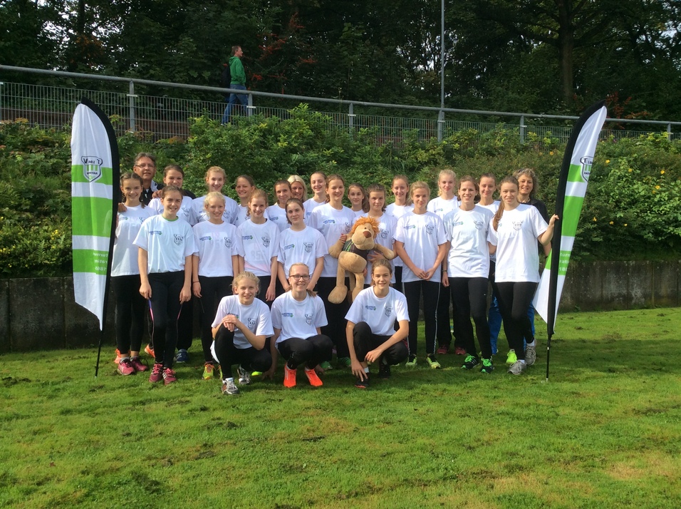 Gruppe von M&auml;dchen in wei&szlig;en T-Shirts mit einem Stoffl&ouml;wen und Fahnen auf gr&uuml;ner Wiese, im Hintergrund B&auml;ume und ein Gehweg.