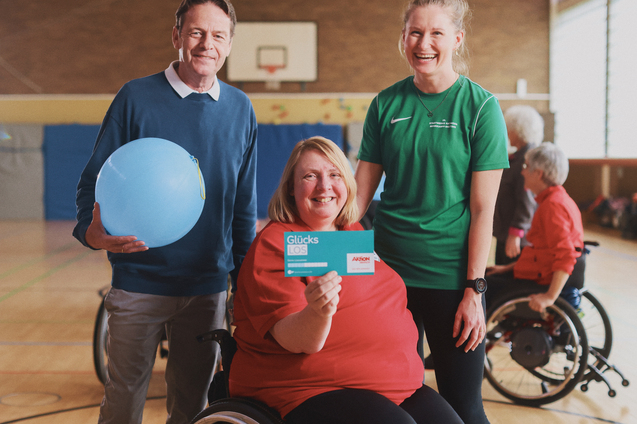 Drei Personen in einem Sportkreis: eine Frau im Rollstuhl h&auml;lt eine Gl&uuml;ckskarte, w&auml;hrend ein Mann und eine Frau l&auml;cheln.