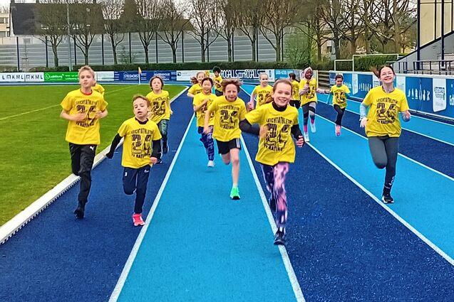 Gruppe von Kindern in gelben T-Shirts l&auml;uft auf einer blauen Laufbahn im Freien. Hintergrund zeigt B&auml;ume und Geb&auml;ude.