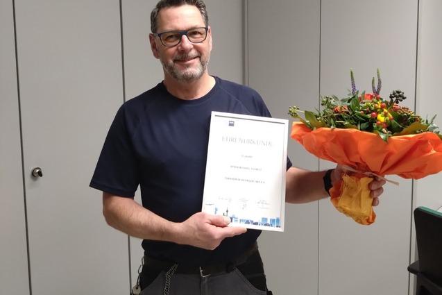 Mann h&auml;lt eine Ehrenurkunde und einen Blumenstrau&szlig; in einem B&uuml;ro mit grauen W&auml;nden und Fliesenboden.
