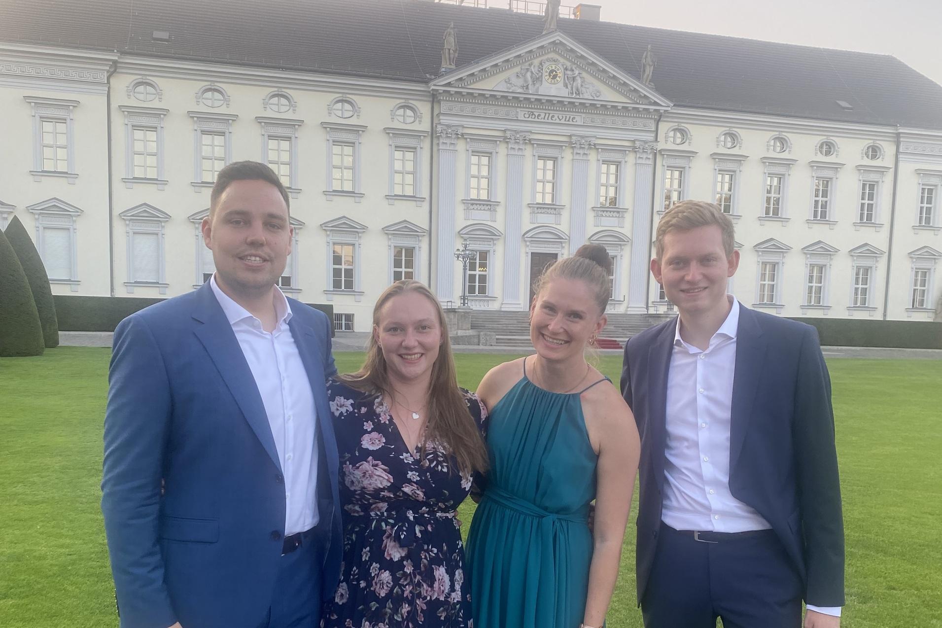 Vier Personen stehen l&auml;chelnd vor einem historischen Geb&auml;ude mit gr&uuml;ner Rasenfl&auml;che und B&auml;umen im Hintergrund.