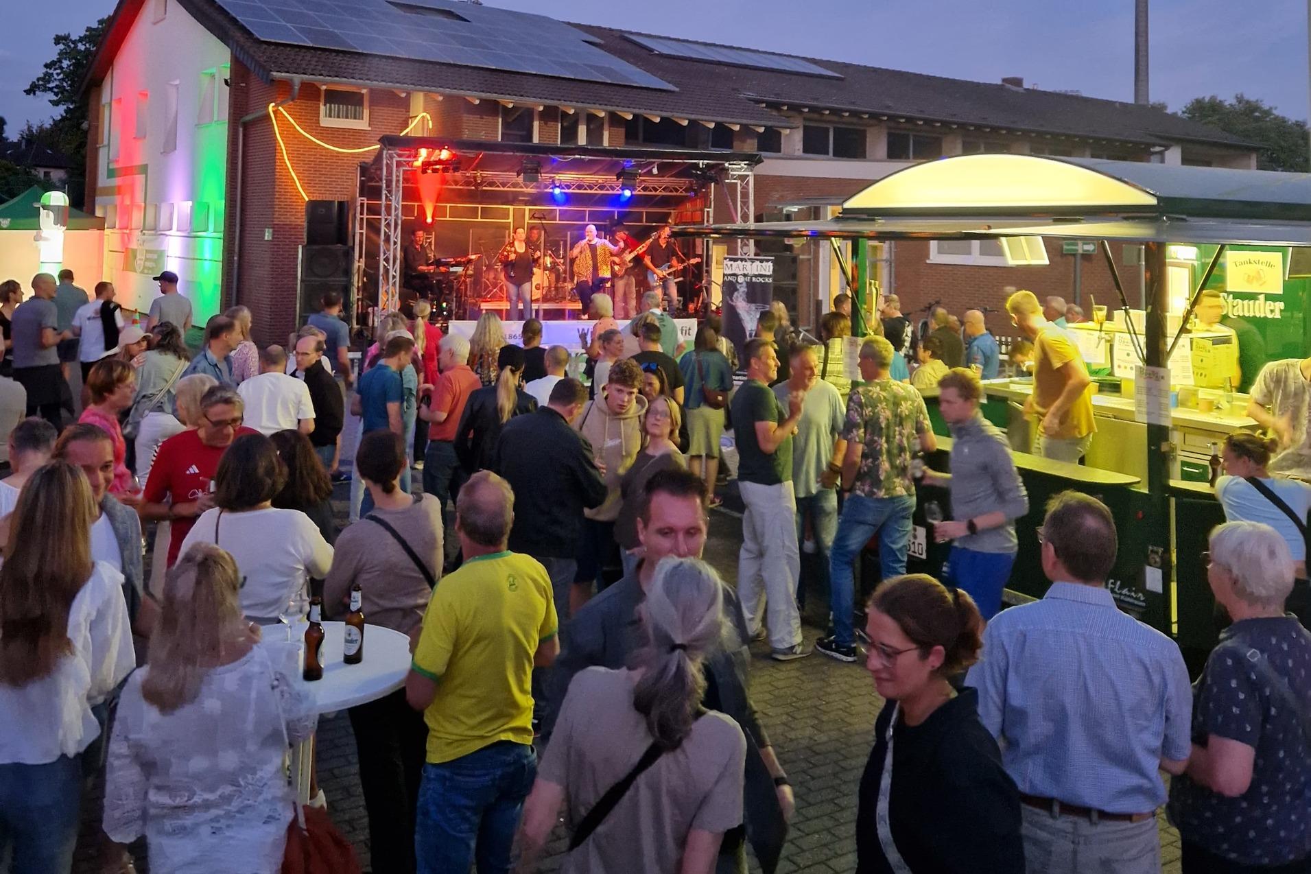 Menschenmenge vor einer Bühne mit Band, umgeben von Ständen und einer beleuchteten Gebäudeansicht bei Dämmerung.