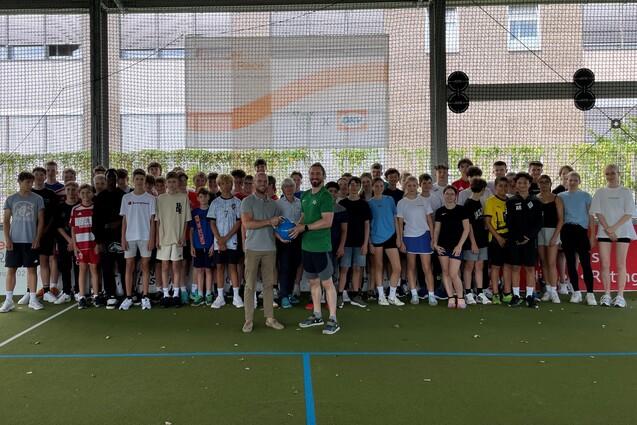 Gruppensch&uuml;ler stehen auf einem Sportplatz, umrahmt von einem Netz, einige Personen halten einen Ball in der Mitte.