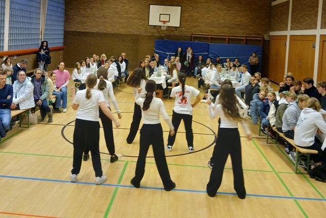 Mehrere T&auml;nzerinnen in Sportbekleidung treten vor einem Publikum in einer Sporthalle auf. B&auml;nke sind im Hintergrund sichtbar.