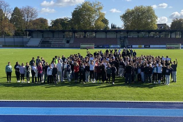 Gruppierung von Menschen auf einem gr&uuml;nen Fu&szlig;ballfeld unter blauem Himmel, im Hintergrund ein Trib&uuml;nenbereich.