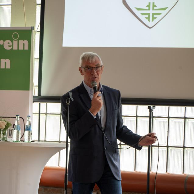 Mann mit Brille spricht am Mikrofon vor einer Präsentation und einem Tisch mit Wasserflaschen und Blumen.