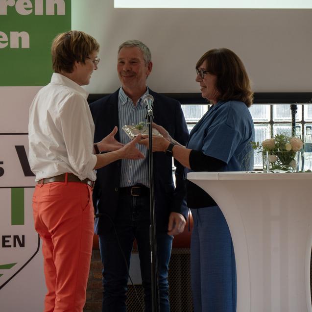 Drei Personen stehen an einem Stehtisch und tauschen eine Auszeichnung während einer Veranstaltung aus.