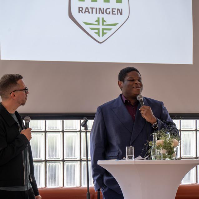 Zwei Männer stehen an einem Stehtisch, der mit Blumen und Getränken dekoriert ist, während im Hintergrund eine Präsentation läuft.