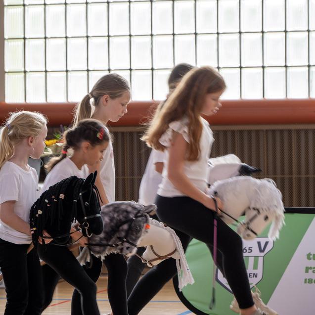 Mädchen mit Spielponys in der Hand laufen auf einem Sportboden, im Hintergrund große Fenster und eine Werbebande.
