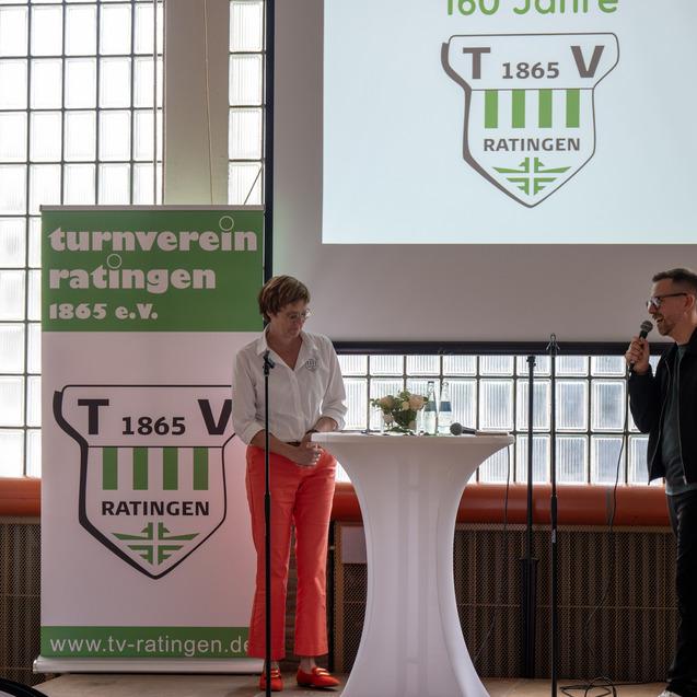 Zwei Personen stehen an einem Stehtisch, während im Hintergrund eine Präsentation des Turnvereins Ratingen 1865 e.V. gezeigt wird.