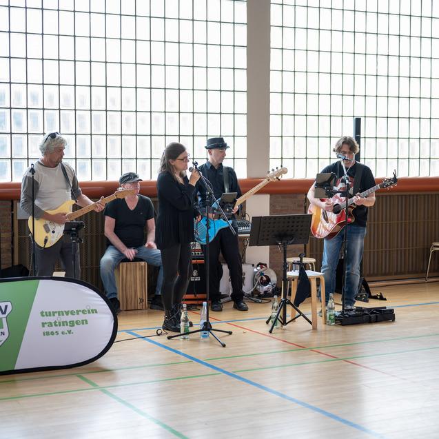 Band mit fünf Musikern spielt in einer Halle, Instrumente sind E-Gitarre, akustische Gitarre und Cajón.
