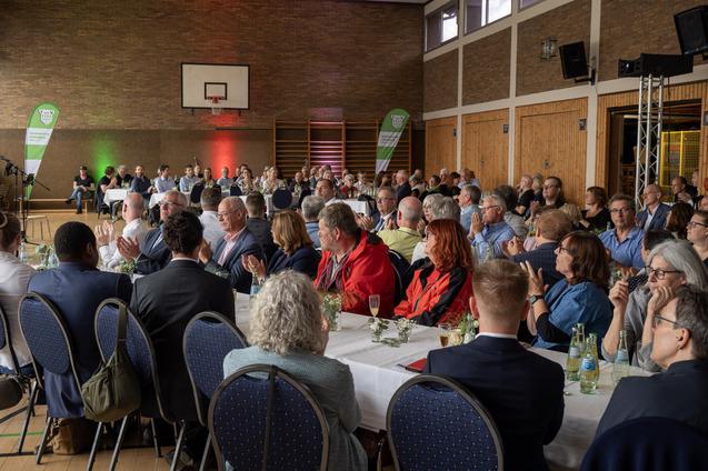 Vielzahl von Menschen in einem festlichen Saal, applaudierend, mit Tischen und dekorierenden Pflanzen im Hintergrund.