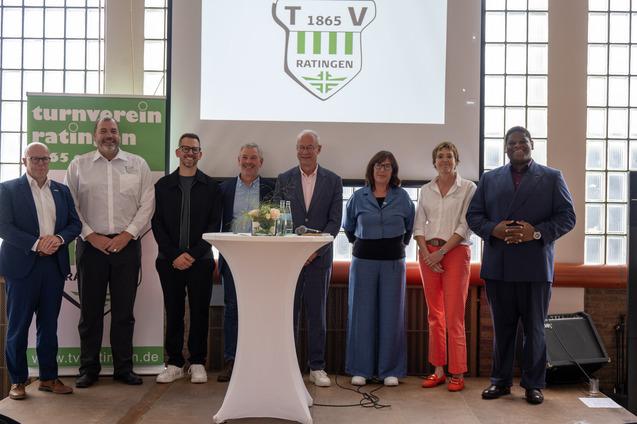 Gruppenfoto von acht Personen vor einer Pr&auml;sentationswand mit dem Logo des Turnvereins Ratingen 1865.