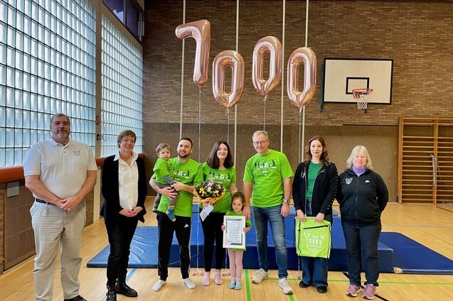 Gruppe von sechs Personen in Sportkleidung mit Luftballons und einer Pflanze in einer Sporthalle.