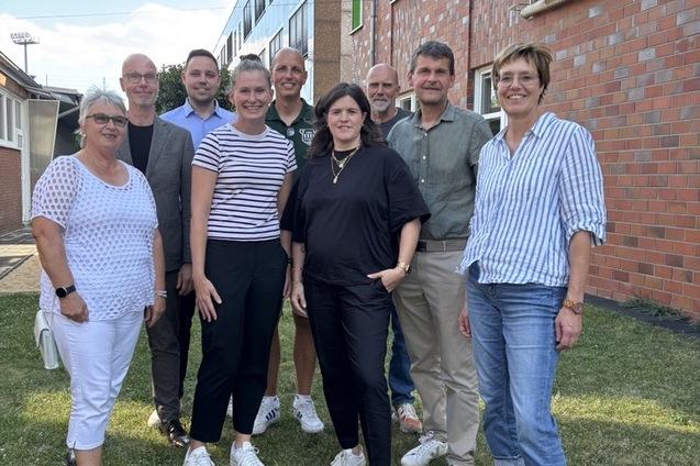 Gruppe von zehn Personen posiert auf einer Wiese, vor einem Backsteingeb&auml;ude an einem sonnigen Tag.