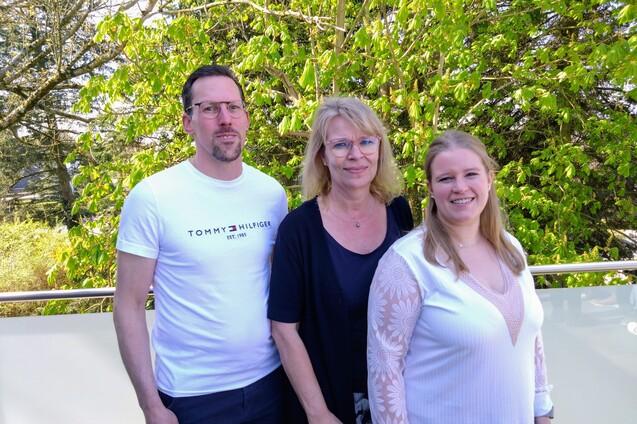 Drei Personen stehen vor gr&uuml;ner Vegetation: ein Mann und zwei Frauen, eine davon l&auml;chelt, die anderen schauen neutral.