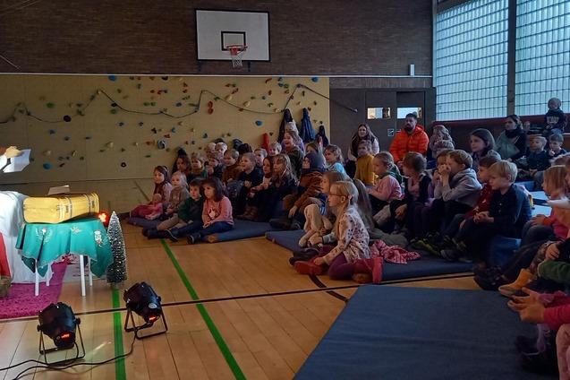 Eine Erz&auml;hlerin in wei&szlig;em Gewand sitzt auf einem Podium vor einer Gruppe von Kindern in einem Hallenraum.
