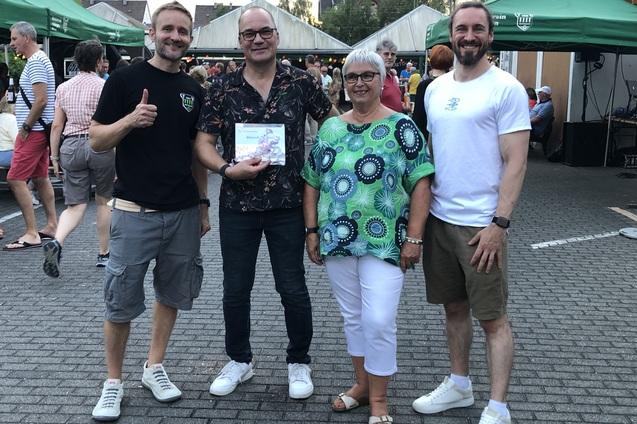 Vier Personen stehen l&auml;chelnd auf einem Fest, eine h&auml;lt eine Auszeichnung in der Hand, im Hintergrund sind Menschen und St&auml;nde.