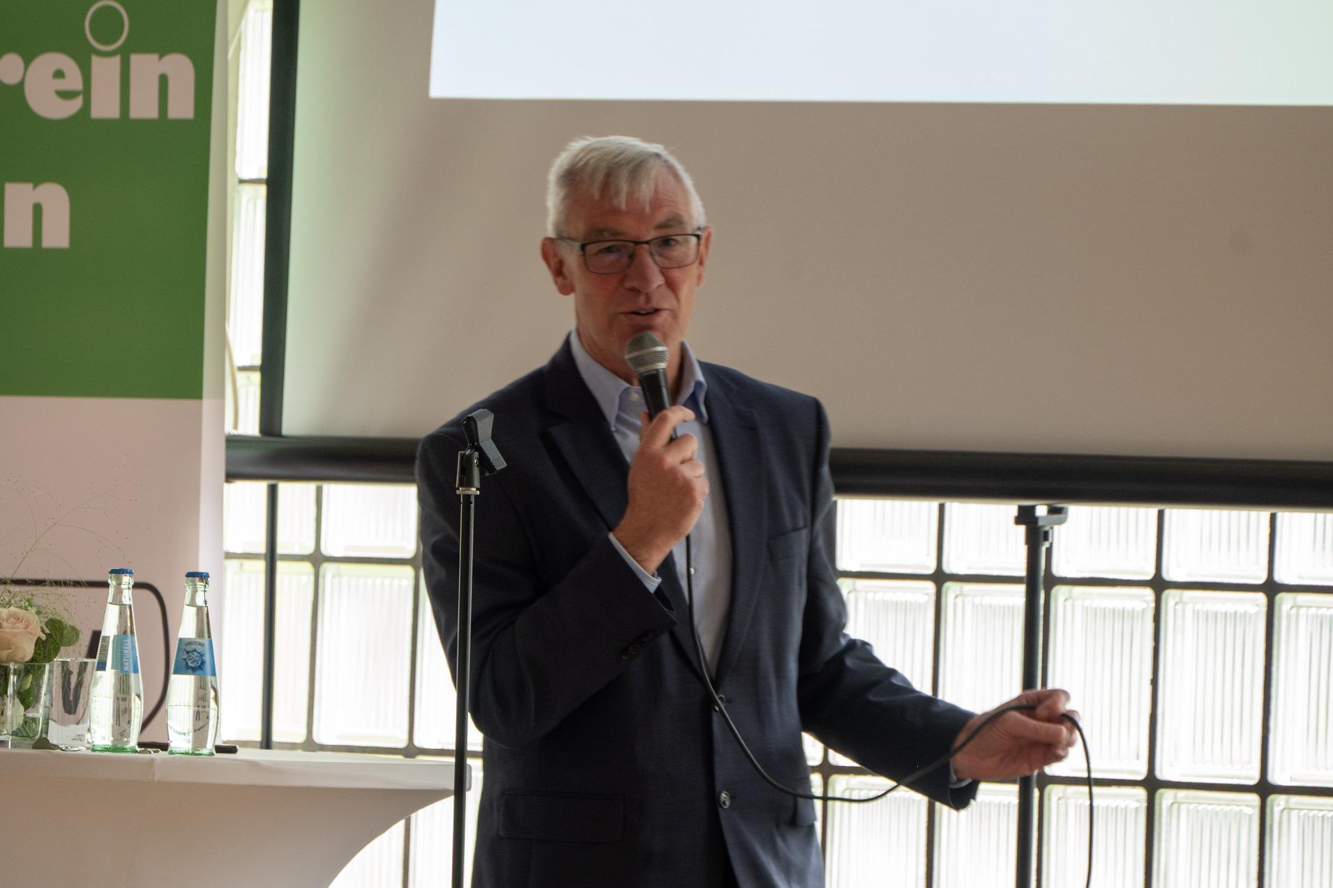 Mann mit Brille spricht am Mikrofon vor einer Präsentation und einem Tisch mit Wasserflaschen und Blumen.