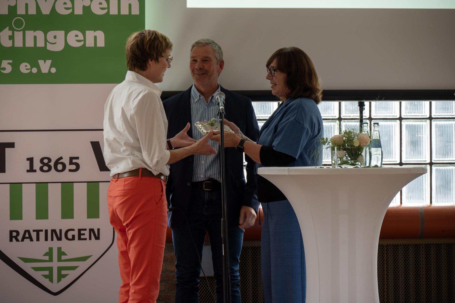 Drei Personen stehen an einem Stehtisch und tauschen eine Auszeichnung während einer Veranstaltung aus.