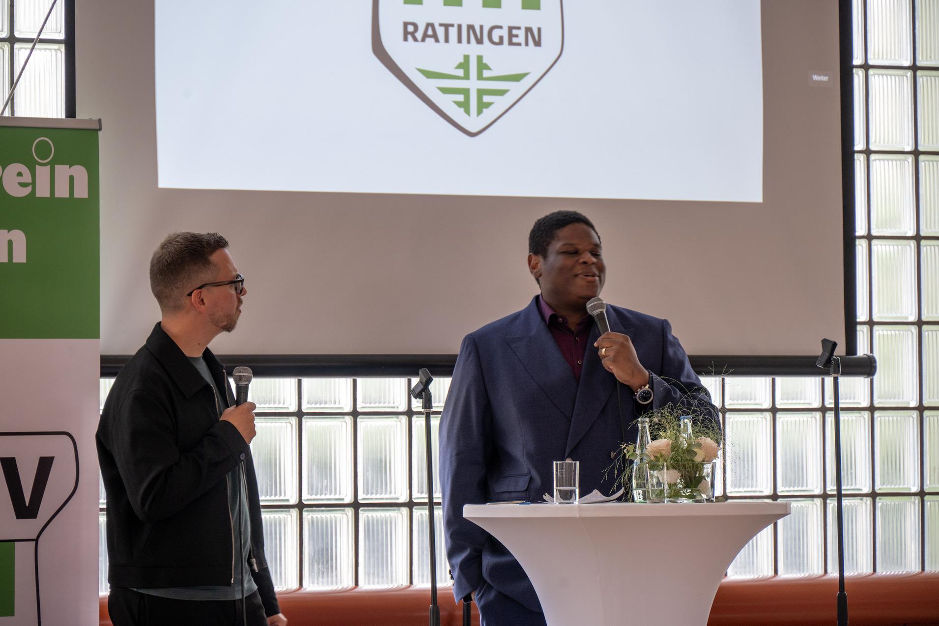 Zwei Männer stehen an einem Stehtisch, der mit Blumen und Getränken dekoriert ist, während im Hintergrund eine Präsentation läuft.