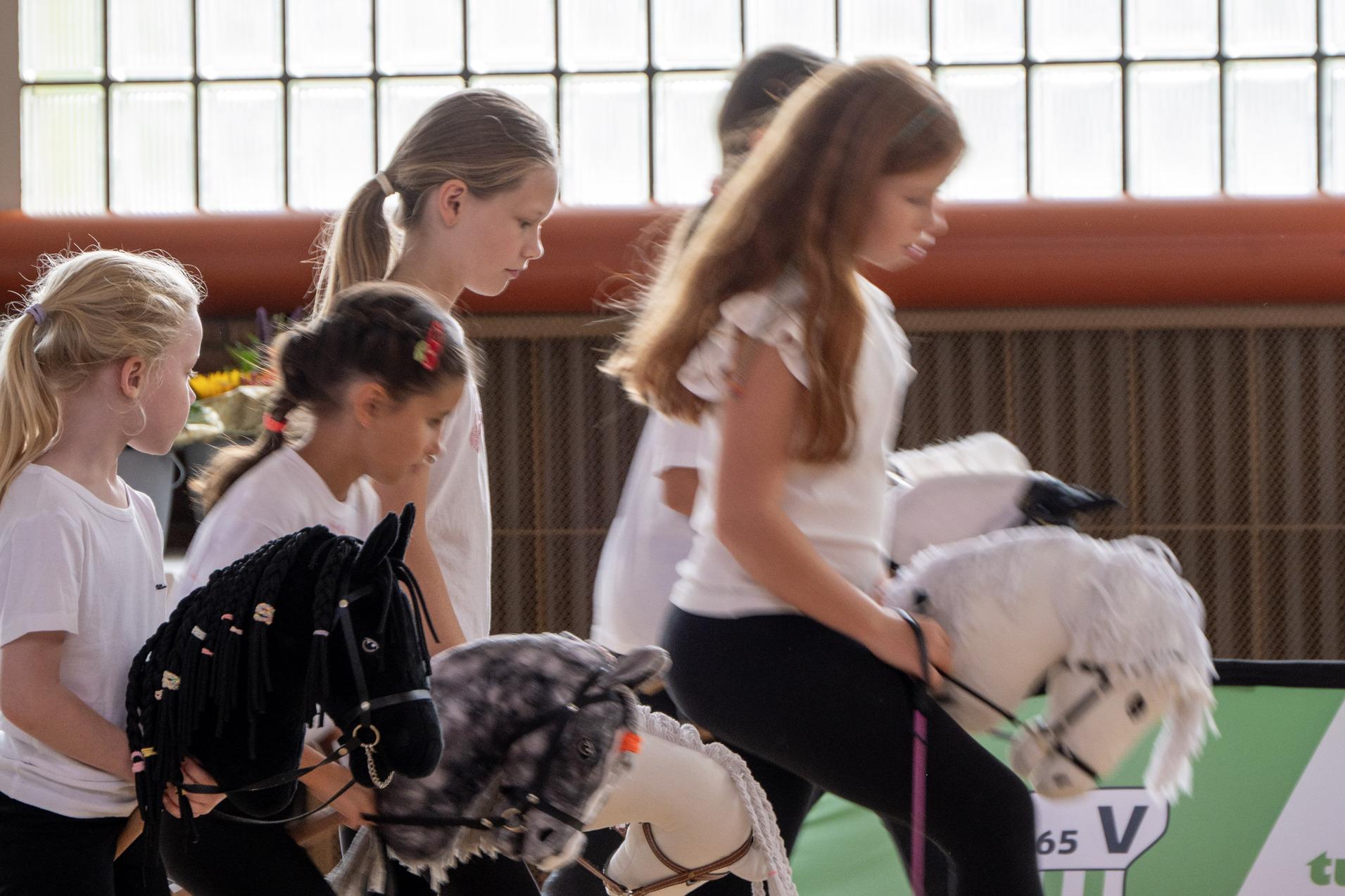 Mädchen mit Spielponys in der Hand laufen auf einem Sportboden, im Hintergrund große Fenster und eine Werbebande.