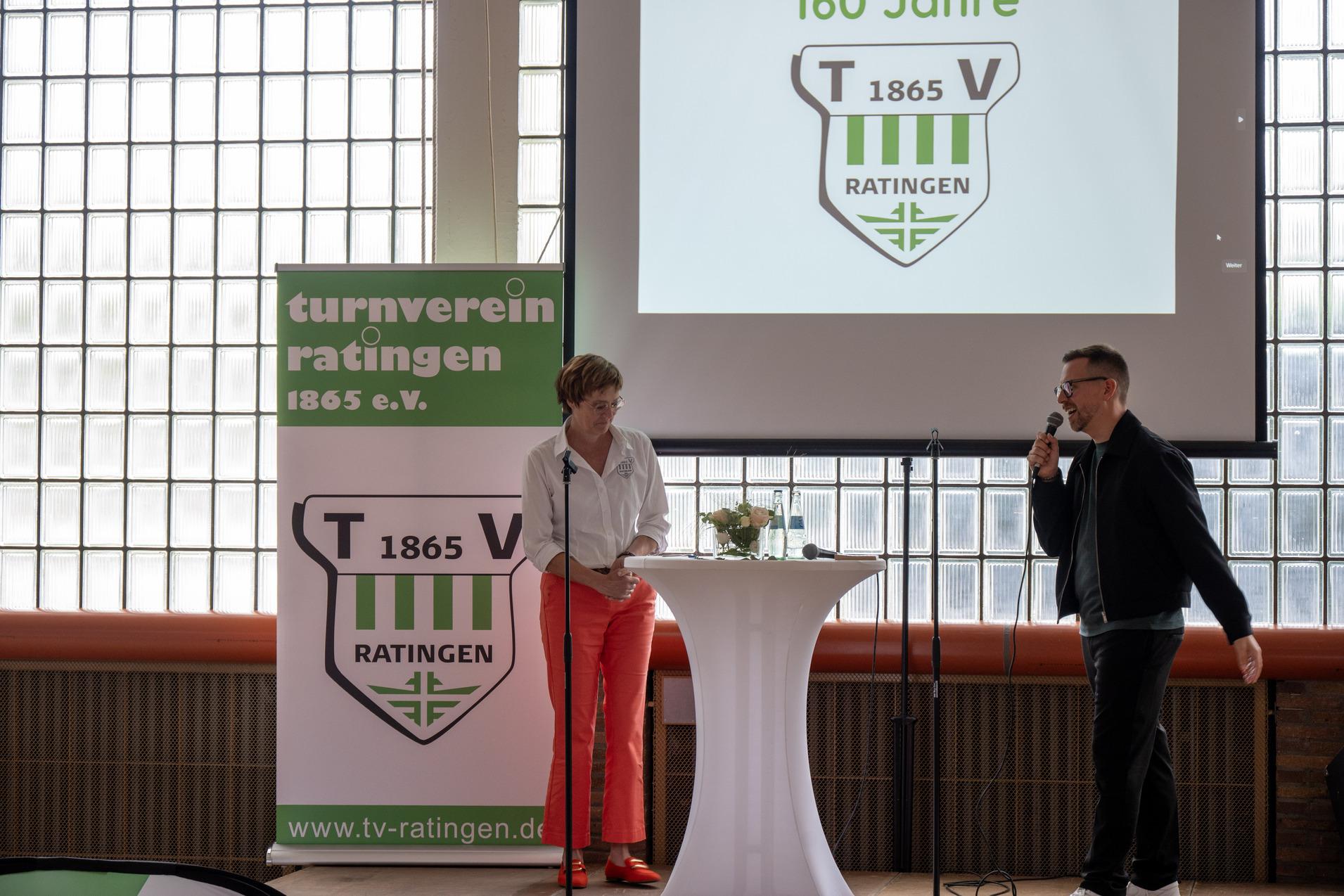 Zwei Personen stehen an einem Stehtisch, während im Hintergrund eine Präsentation des Turnvereins Ratingen 1865 e.V. gezeigt wird.
