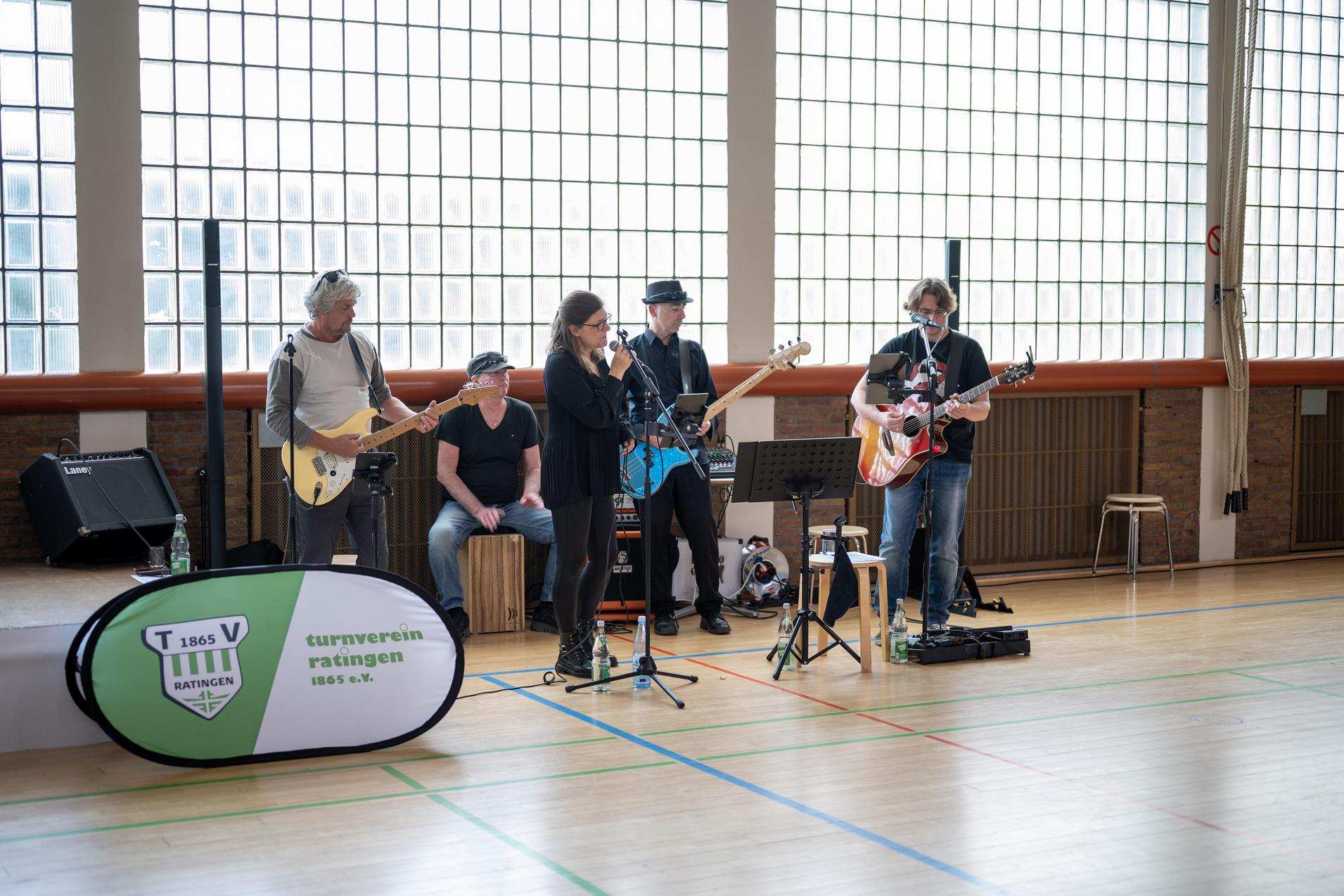 Band mit fünf Musikern spielt in einer Halle, Instrumente sind E-Gitarre, akustische Gitarre und Cajón.