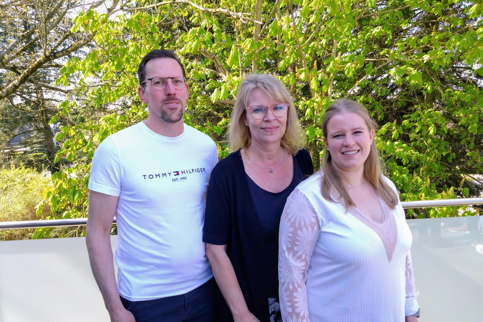 Drei Personen stehen vor gr&uuml;ner Vegetation: ein Mann und zwei Frauen, eine davon l&auml;chelt, die anderen schauen neutral.