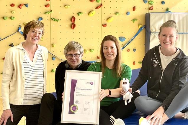 Vier Personen sitzen auf einem blauen Turnger&auml;t, halten eine Urkunde und einen wei&szlig;en Stoffhasen. Kletterwand im Hintergrund.