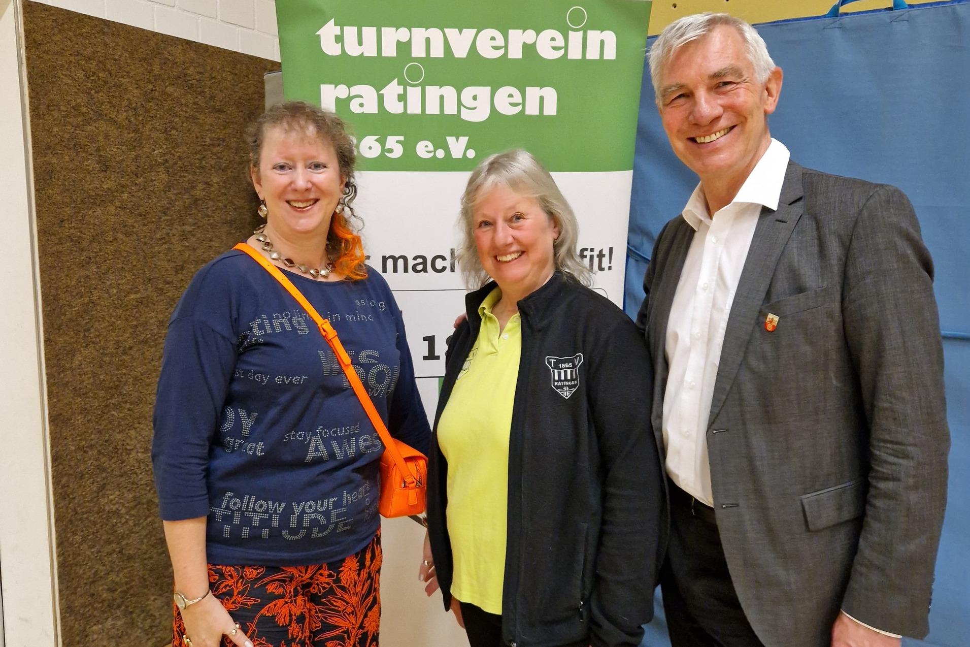 Drei Personen stehen l&auml;chelnd vor einem Banner des Turnvereins Ratingen in einer Sporthalle.
