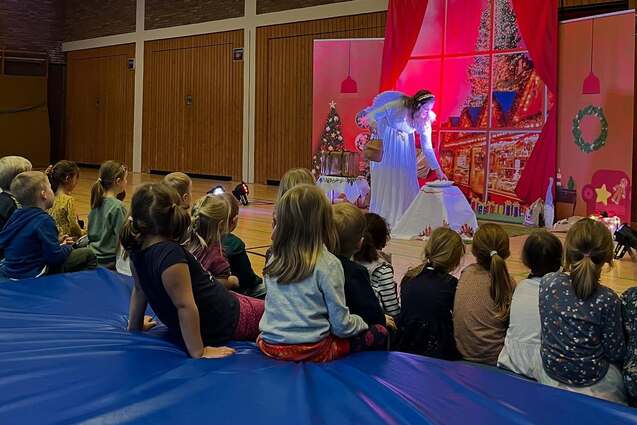 Eine junge Performer/in in wei&szlig;em Kost&uuml;m f&uuml;hrt vor einer Gruppe Kinder auf einer B&uuml;hne mit weihnachtlicher Dekoration auf.