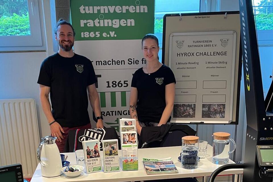 Zwei Personen stehen an einem Tisch mit Informationsmaterialien zur HYROX Challenge in einem Sportzentrum.