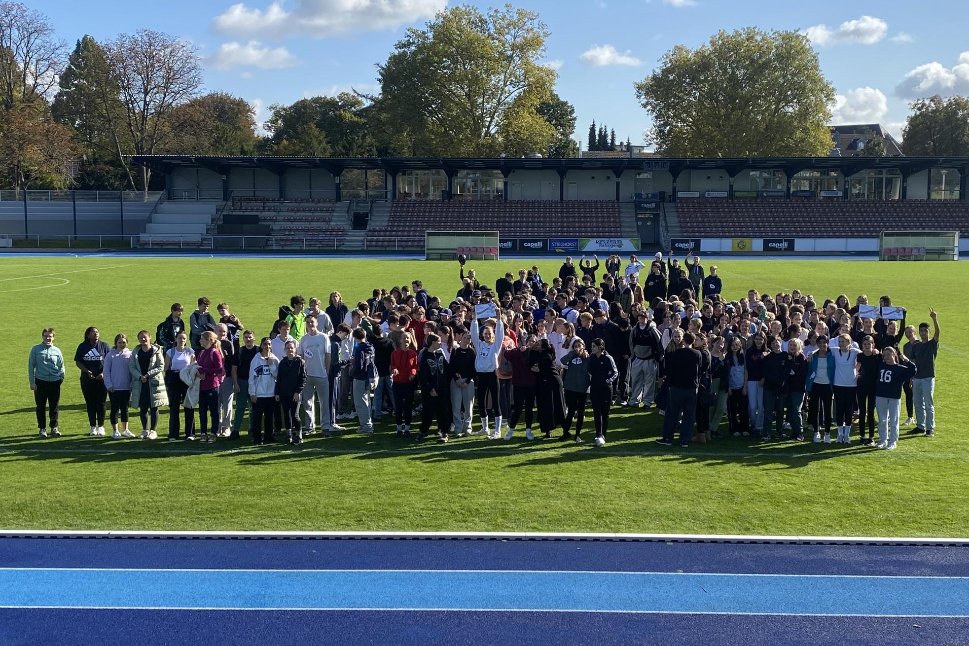 Gruppierung von Menschen auf einem grünen Fußballfeld unter blauem Himmel, im Hintergrund ein Tribünenbereich.