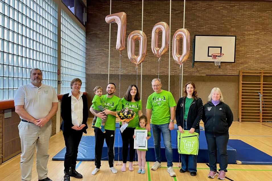 Gruppe von sechs Personen in Sportkleidung mit Luftballons und einer Pflanze in einer Sporthalle.