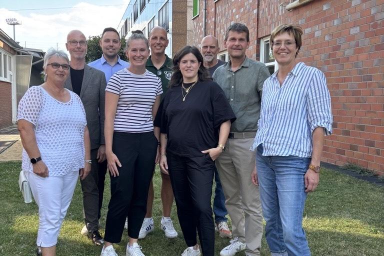 Gruppe von zehn Personen posiert auf einer Wiese, vor einem Backsteingebäude an einem sonnigen Tag.
